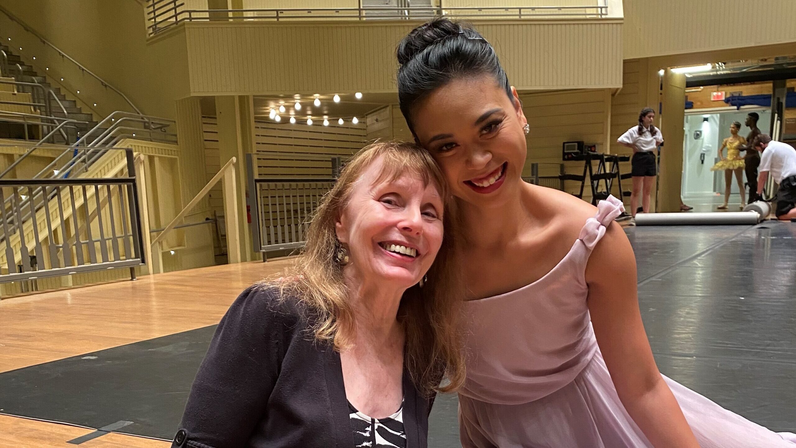 Patricia McBride and Angelica Generosa pose and smile, their heads leaning against each other. Genrosa wears a pink filmy dress, while McBride wears a short black jacket and white and black patterned dress.