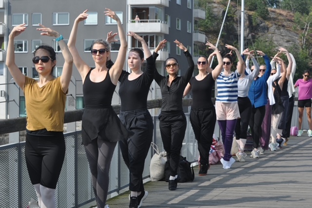 A long line of adult dancers wearing practice clothes and sunglasses pose in suos-sus as they take ballet class outdoors, using a bridge as their barre.