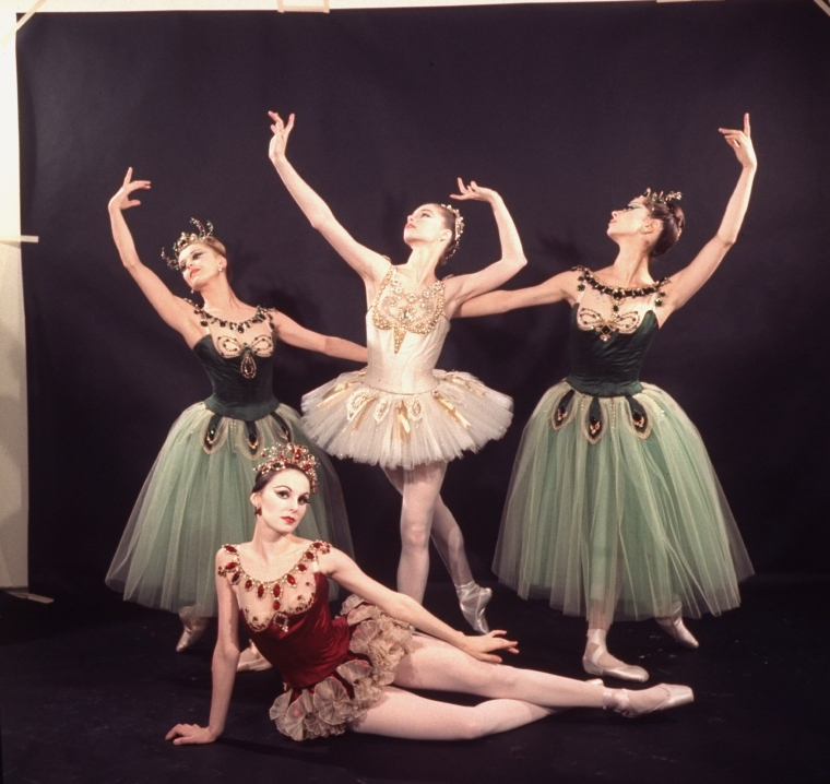 Four ballerinas pose for a photo against a black backdrop. One is seated sideways in front, propped up on her right hand with her legs extended; she wears a short red tutu with red jewel embellishments. Behind her, a ballerina in a short white tutu stands between two in long green tutus in b plus.