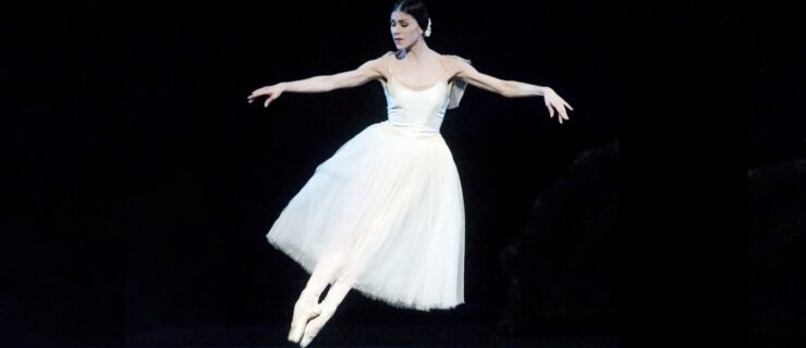 Paloma Herrera, wearing a white Romantic tutu and pointe shoes, does an assemblé with her arms in demi-second position. She looks down over her feet, and looks as if she's hovering above the ground.
