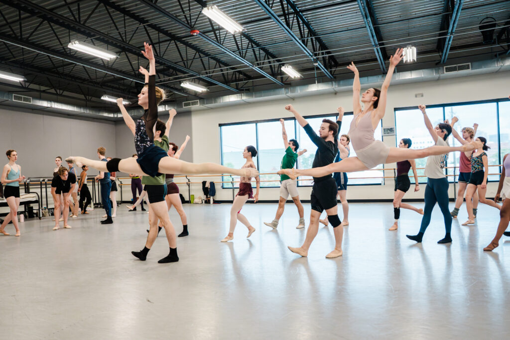 Two female ballet dancers in practice clothes do a saut de chat with their arms lifted in a V position. A large group of male and female dancers run behind them in a large dance studio.