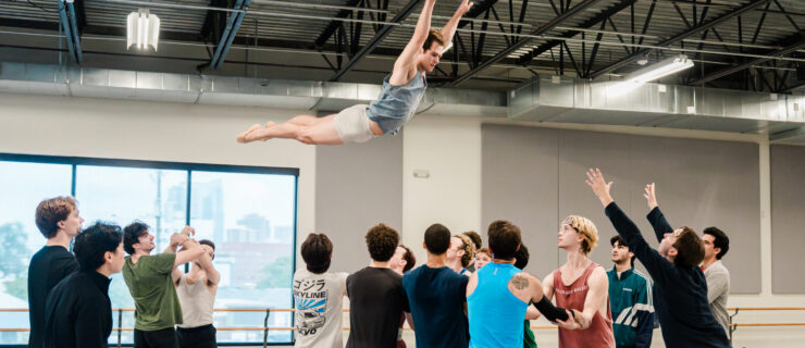 A male dancer in shorts and a t-shirt flies high in the air, arms over his head, as a group of other dancers stand below him with arms linked, ready to catch him. A man at the end reaches his arms up towards the dancer in the air.