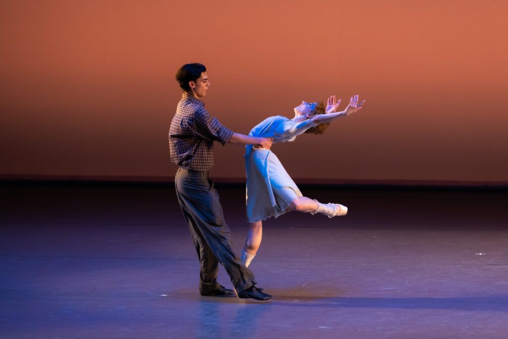 Two teenage dancers performa a pas de deux onstage. The girl, wearing a light blue dress and tattered socks, does a piqué attitude on pointe, arching back with her arms raised. She faces her partner, who is dressed in gray pants and a brown shirt and holds her by the waist.