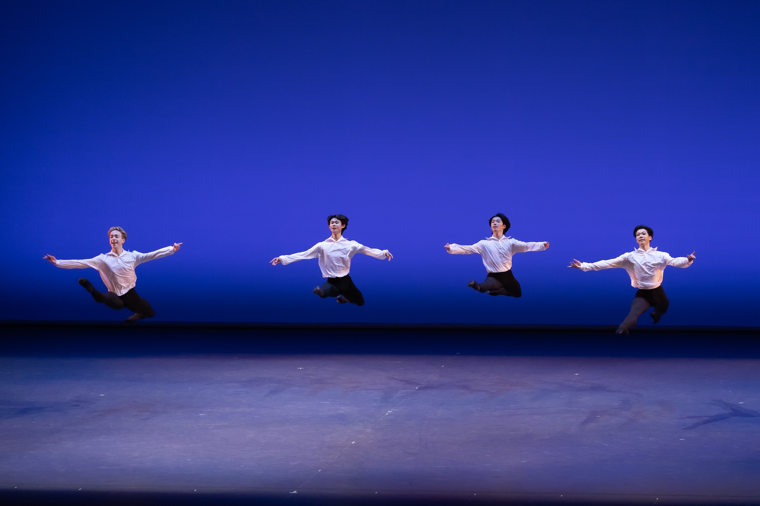 Four male dancers in billowy white shirts, lack tights, and black ballet slippers do a grand jeté in a line coming downstage.