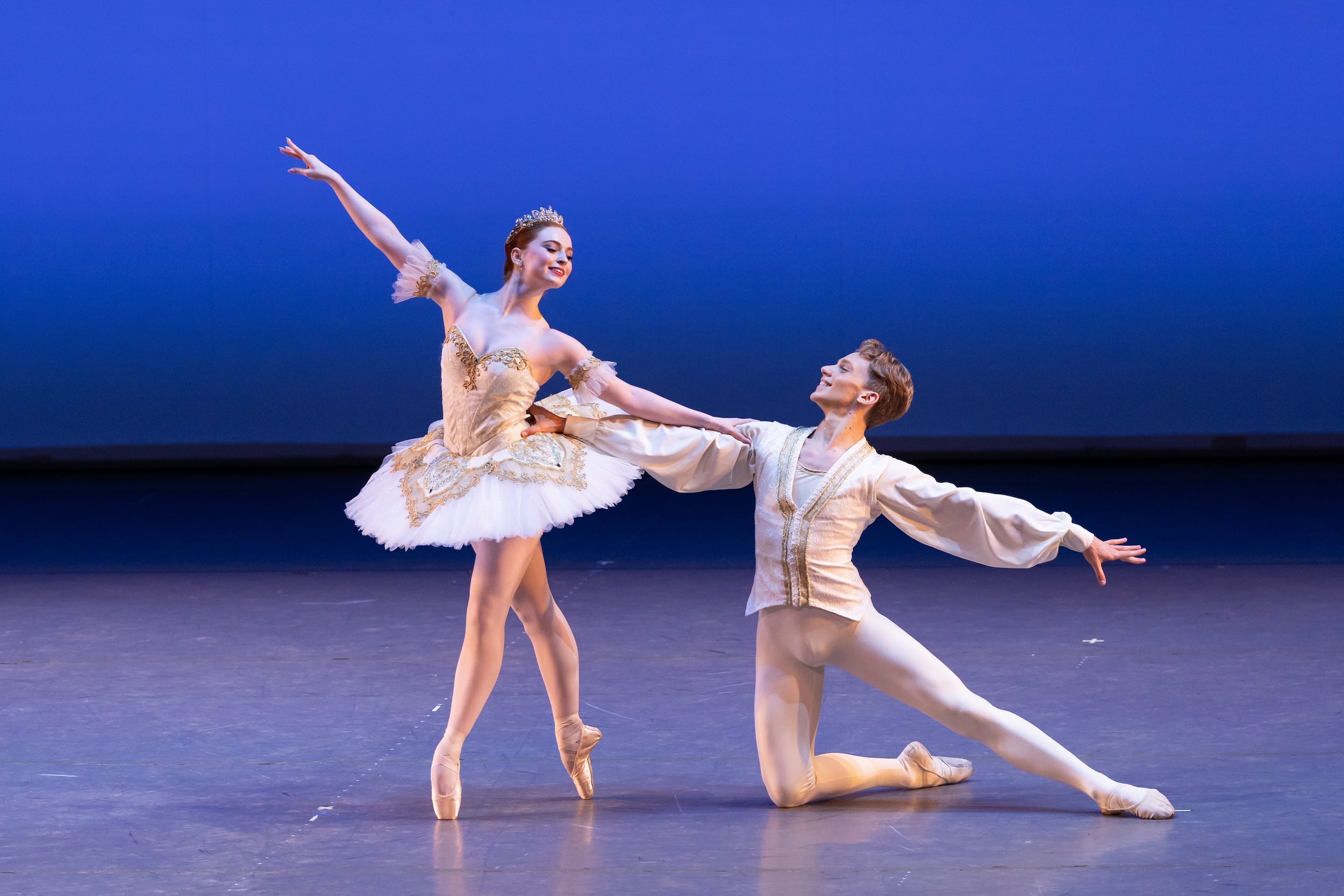 Two teenage dancers pose while performing a pas de deux onstage. The girl, in a white tutu, stands in fourth position croisé on ponte and looks back at her partner, costumed in a white tunic and tights, as he kneels to her left and holds her by the waist.