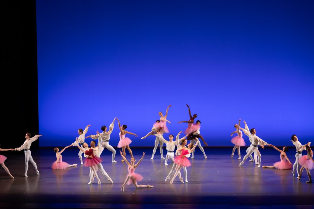 A large group of teenage ballet dancers perform a classical ballet onstage. The women wear pink tutus and the men are in white shirts and tights. They form an inverted V-shape and stand in vairous lifts and poses, while two couples and a soloist pose downstage center.