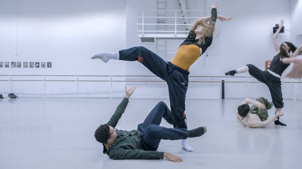 Two pairs of dancers rehearse in a large ballet studio. The women lean back on one leg with the other lifted and bent. They cross their arms above and look up. Beneath them, their partners lie on their backs with bent legs off the floor, reaching up with one arm as if to shield themselves.