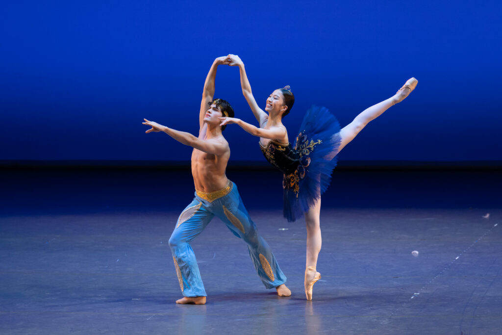 Two young ballet dancers perform a pas de deux onstage. A boy in blue harem pants lunges forward as a girl in a blue tutu does piqué arabesque on pointe, holding on to his raised right hand as they both extend their left arms forward.