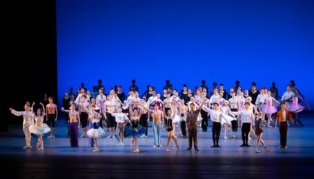 A large group of teenage ballet dancers in various costumes take a bow onstage.