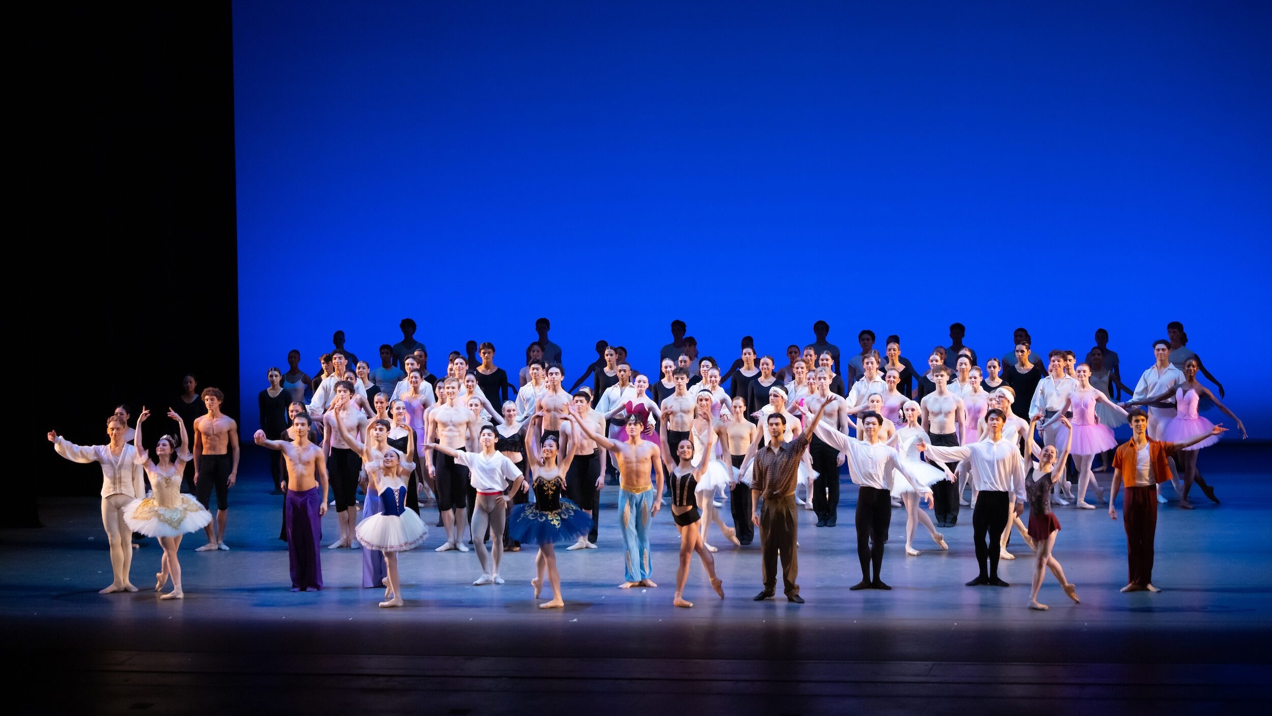 A large group of teenage ballet dancers in various costumes take a bow onstage.