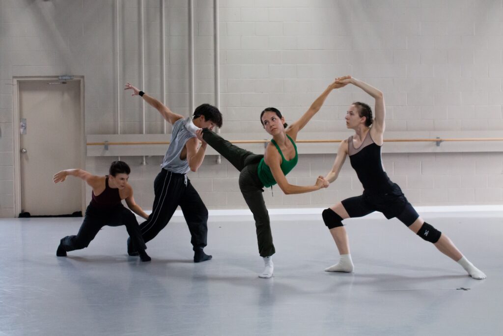 In a brightly lit ballet studio, four dancers form a line while doing various contemporary ballet poses. They each touch the person in front of them, holding a leg, hand, or calf.
