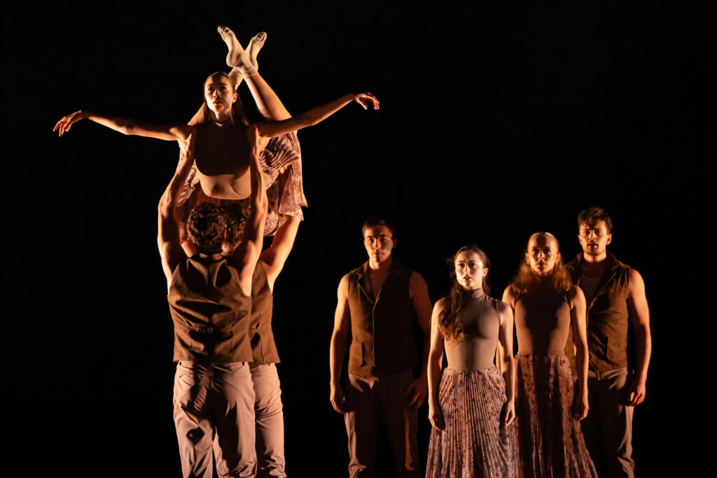 Onstage with dark lighting, seven dancers perform a contemporary ballet wearing neutral-colored costumes. In front, two men lift a woman abovehead as she arches her back and crosses her ankles with knees bent, reaching her arms out to either side. The other four dancers stand behind them, slightly right, with arms straight down.