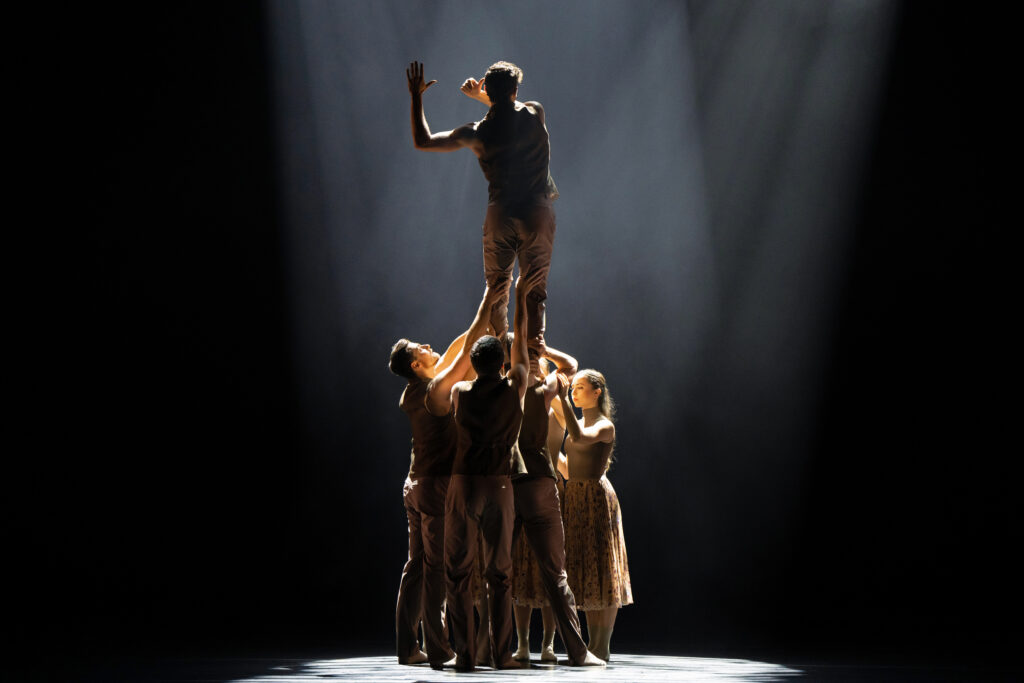 Onstage in dark lighting, a group of dancers in neutral-colored costumes cluster center stage in a spotlight as they lift up one man. The man faces back, one arm lifted with a bent elbow, as he crosses the other in front of his body with a slight twist.