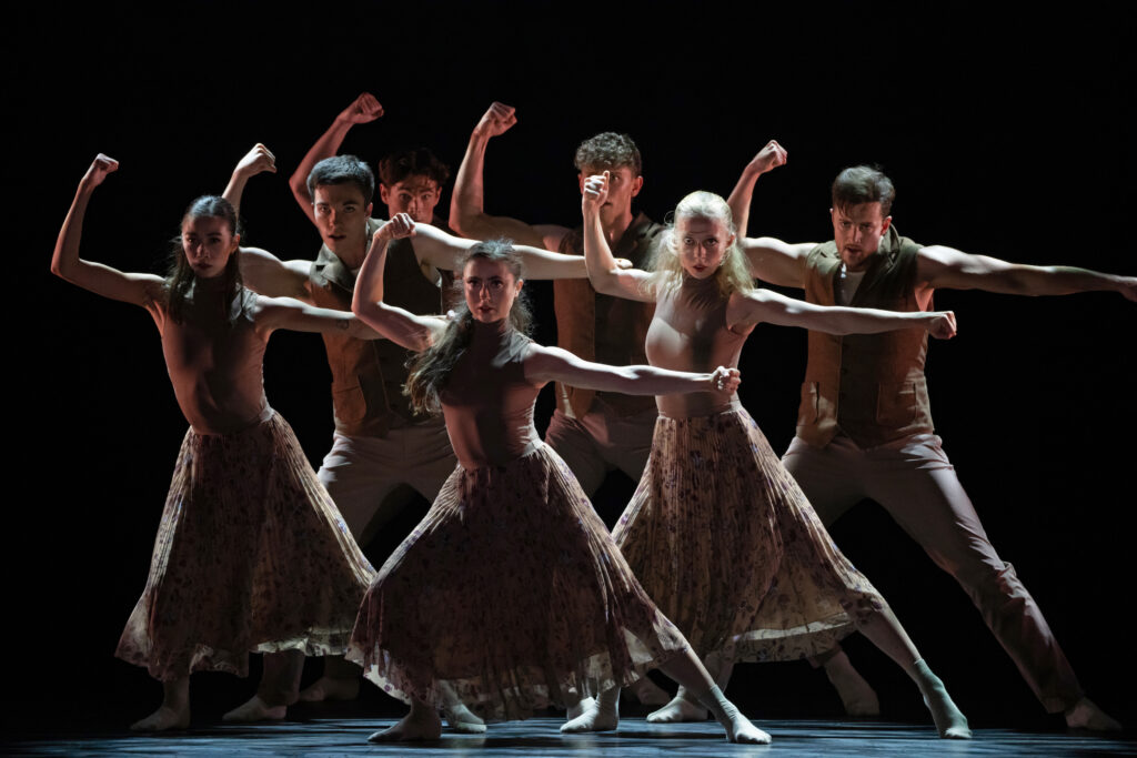 Onstage in dark lighting, a group of dancers in neutral-colored costumes stand in a clump and perform the same motion. They shift their weight sideways with one leg bent, one arm bent straight up at the elbow with the other extended sideways, both hands in fists.