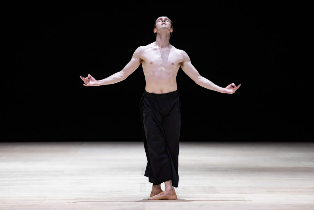 Callum Linnane, wearing loose black pants and ballet shoes, stands onstage in a tight fifth position. He extends his arms to either side, slightly curved up, with his pointer fingers bent to touch his thumbs. He closes his eyes and tilts his chin upward with emotion.
