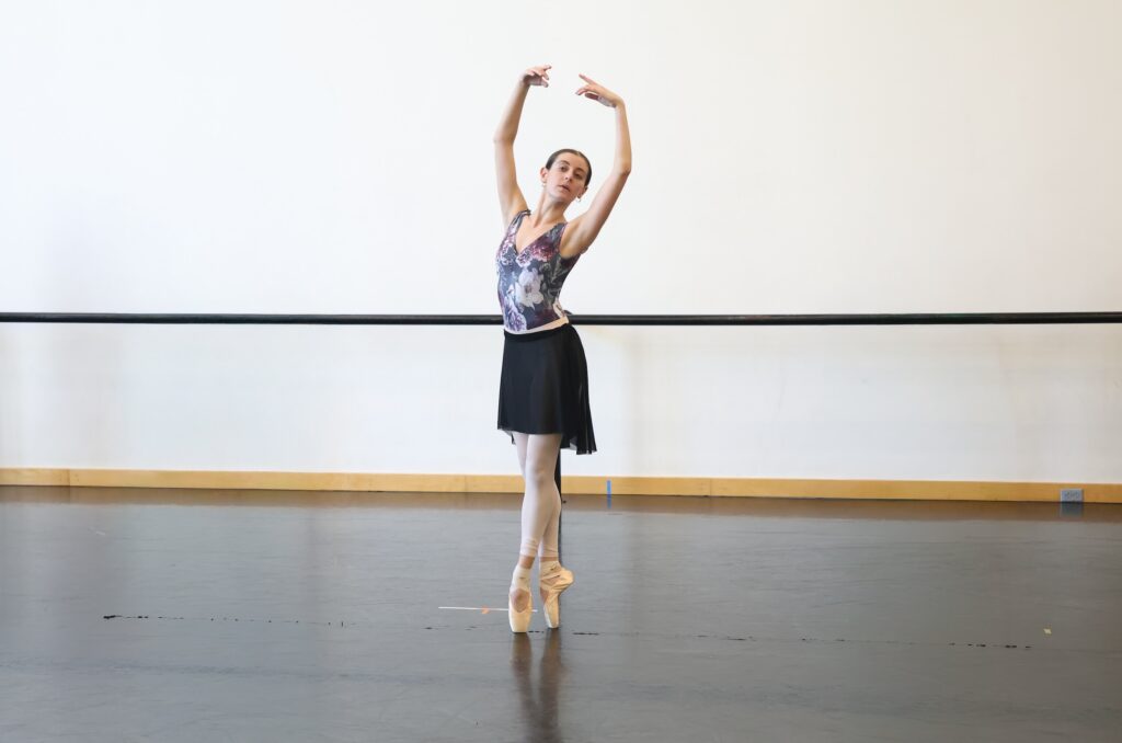 Atherna King, wearing a floral patterned leotard, black ballet skirt, pink tights rolled up at the ankle, stands in sous-sus on pointe with her arms in high fifth.