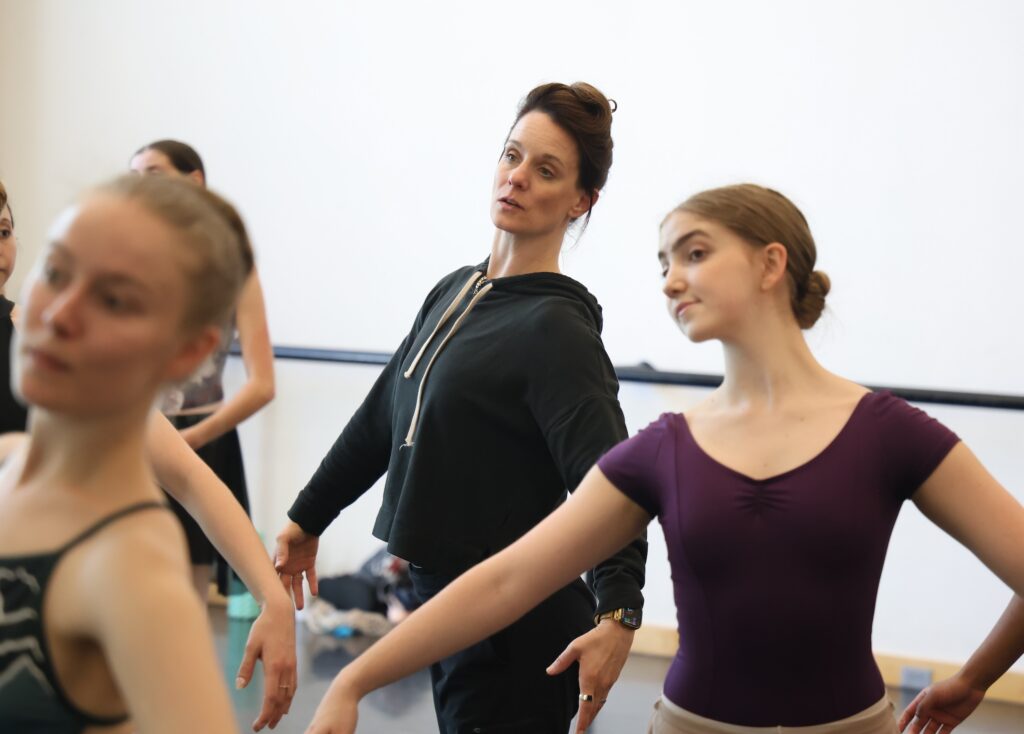 Jill Krutzkamp wears a dark sweatshirt and pants and stands behind a group of female ballet students in practice clothes during a ballet class. They all hold their arms in demi-second position, using epaulment.