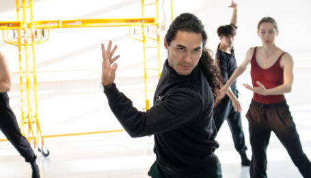 Cameron sinkʷə Fraser-Monroe, shown from the hips-up, twists his upper body with one arm crossed in front, bent at the elbow with his fingers reaching upward. He looks into the mirror (off-camera) with intensity. Ballet dancers behind him watch and mimic his movement.