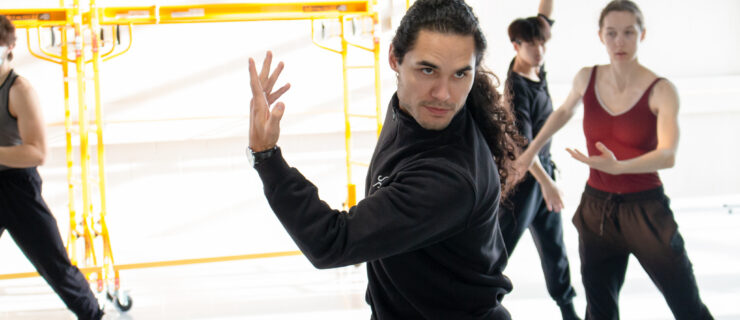 Cameron sinkʷə Fraser-Monroe, shown from the hips-up, twists his upper body with one arm crossed in front, bent at the elbow with his fingers reaching upward. He looks into the mirror (off-camera) with intensity. Ballet dancers behind him watch and mimic his movement.