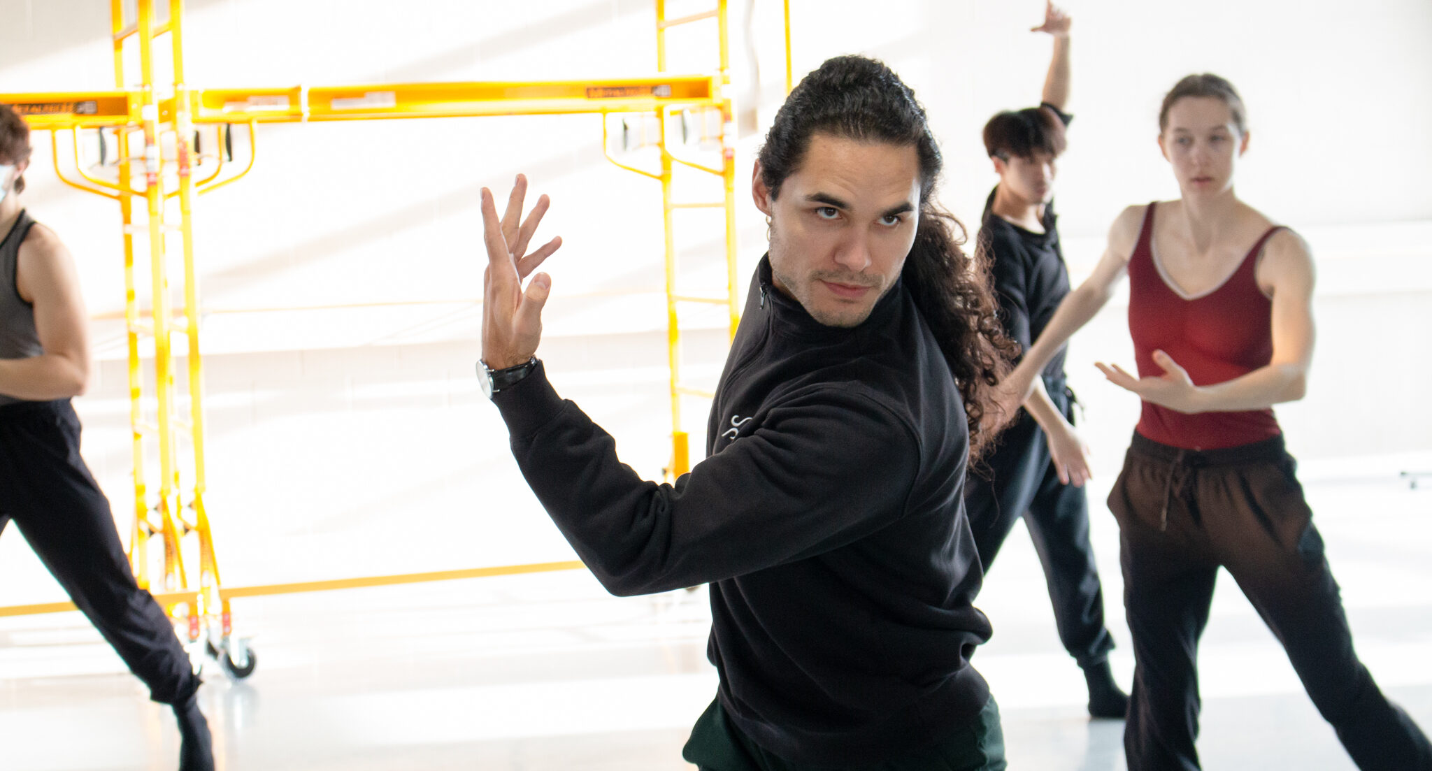 Cameron sinkʷə Fraser-Monroe, shown from the hips-up, twists his upper body with one arm crossed in front, bent at the elbow with his fingers reaching upward. He looks into the mirror (off-camera) with intensity. Ballet dancers behind him watch and mimic his movement.