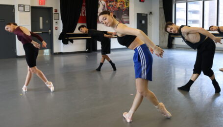 In a ballet studio, an ensemble of dancers pose with one leg in b-plus, toes flexed, with their upper bodies curved forward. They curve their arms up and back to match the line and tilt their heads to the side.