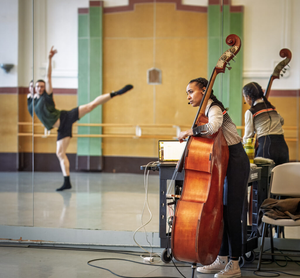 In a ballet studio, esperanza spalding plays her bass while watching a dancer intently, whose reflection in the mirror shows him in a fondu arabesque with one arm high.