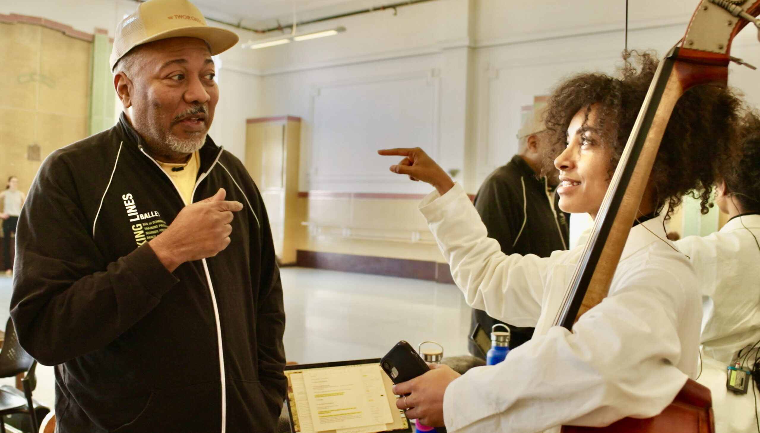 In a ballet studio, Alonzo King and esperanza spalding converse in front of a large mirror with a laptop open behind. They both lift an arm as they speak, and spalding cradles a bass in the crook of one arm.