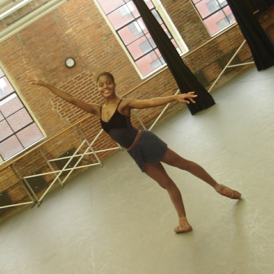 A teenage Ingrid Silva stands in tendu derrière with her arms in first arabesque position. She wears a dark leotard, brown tights and pointe shoes, and a dark ballet skirt, and poses in a dance studio in front of a brick wall