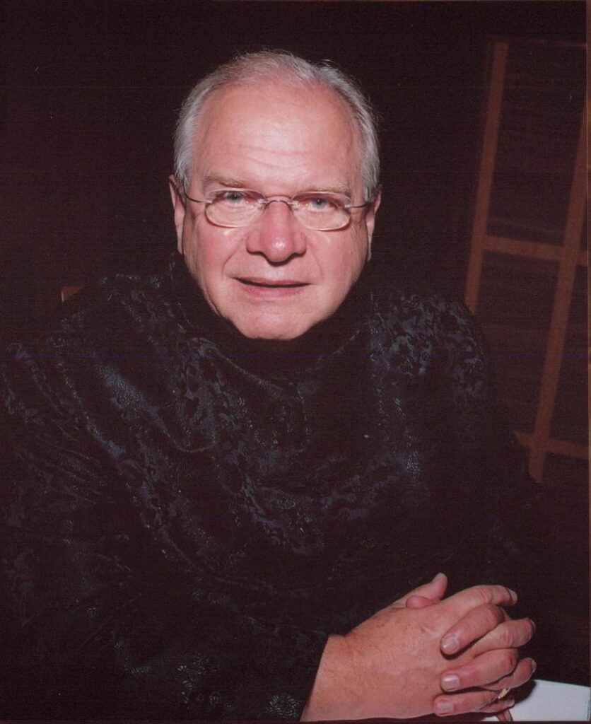 Ben Stevenson's headshot. He wears a black suit with crushed velvet and wire-rimmed glasses.