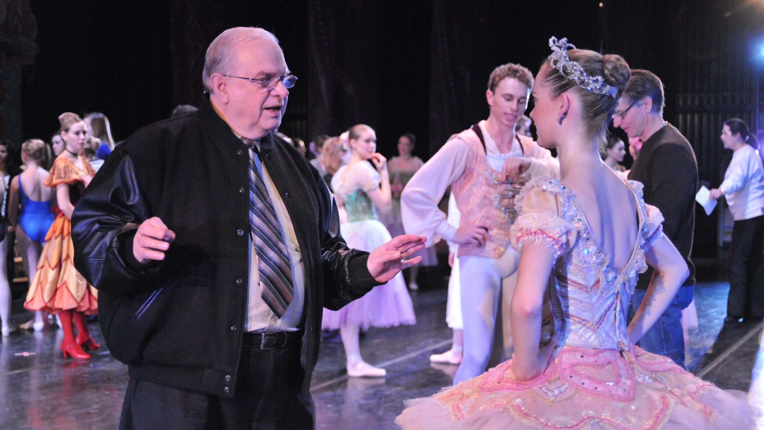 Ben Stevenson, wearing black pants and a black jacket over a white shirt and striped tie, talks to a ballerina in a pink tutu backstage. Other dancers in costume mill about in the background.
