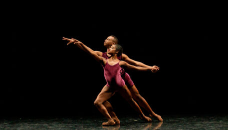 David Wright stands directly behind Ingrid Silva as they lunge forward on their right leg toward stage left, holding left hands. They both wear purple leotards and perform onstage in front of a black backdrop.