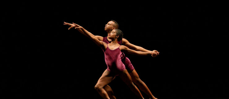 David Wright stands directly behind Ingrid Silva as they lunge forward on their right leg toward stage left, holding left hands. They both wear purple leotards and perform onstage in front of a black backdrop.
