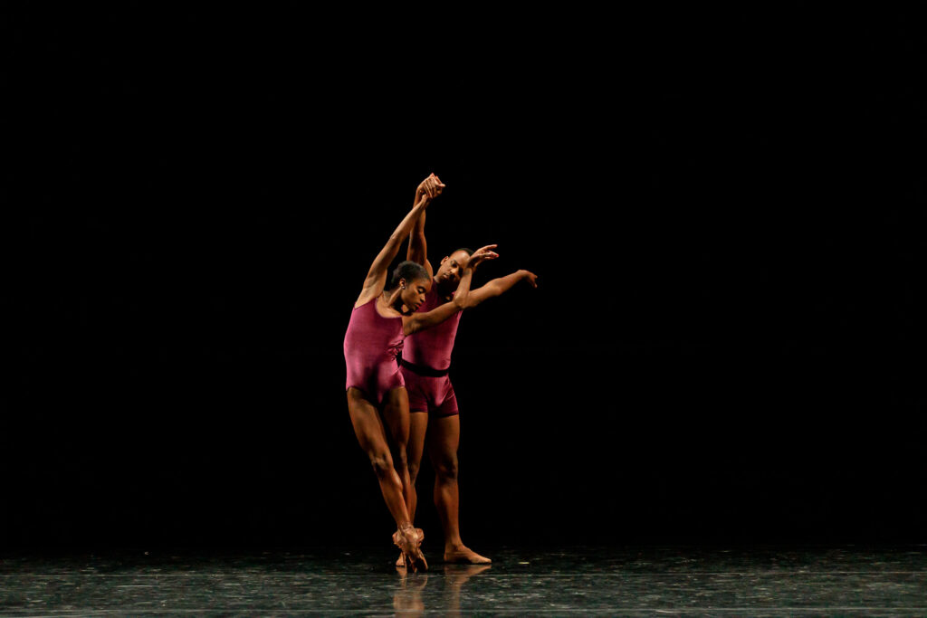 Ingrid Silva and David Wright dance a pas de deux onstage in front of a black backdrop. Silva, in a purple leotard, stands in sus-sous, leaning slightly to her right, as Wright, in a purple biketard, stands behind her and holds her raised right arm.