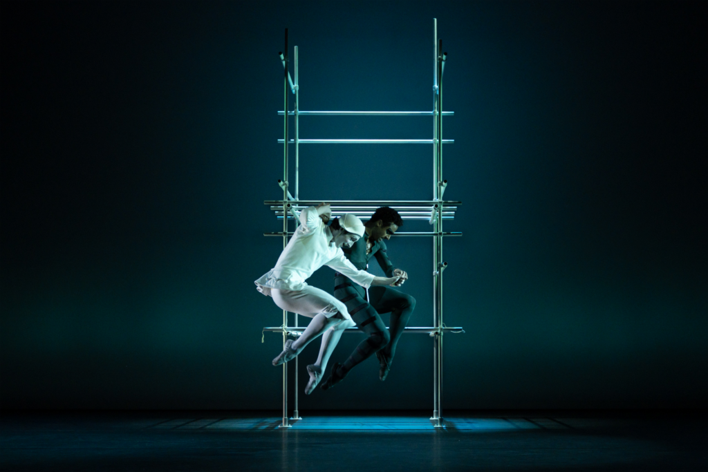 Two dancers jump up in front of metal scaffolding onstage, their legs tucked under and their bodies in profile facing stage left.. The dancer on the left is dressed as a clown, wearing a white cap and top, matching pantaloons, white tights, and white shoes, with his face painted white. The other dancer is behind him, and wears a dark green jacket, knickers, tights and shoes.