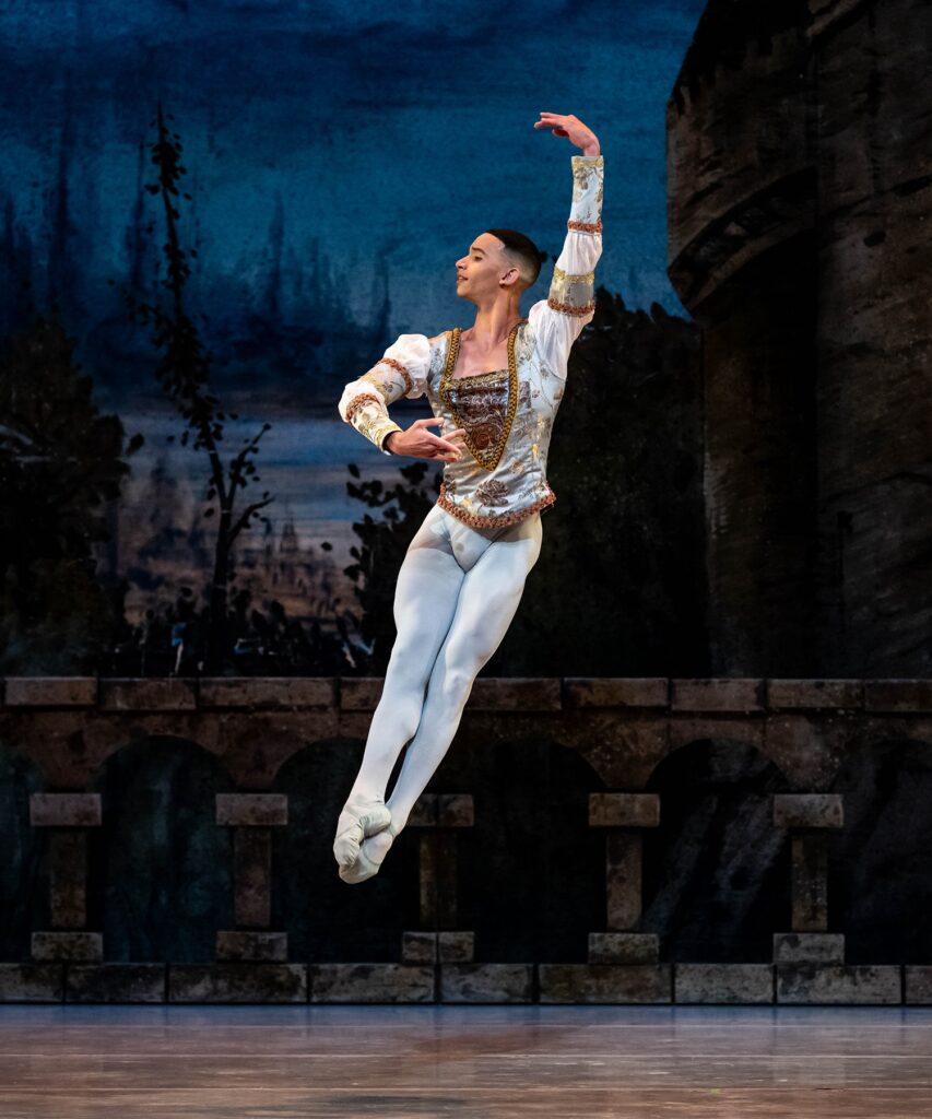 Randolph Fernandez, wearing white tights, white ballet clippers, and a white tunic with gold trim, does an assemblé onstage. He holds his left arm up and his right arm in front. of him and looks over his right shoulder, smiling.