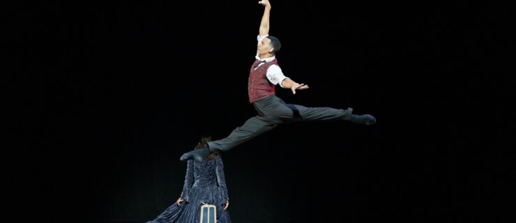Randolph Fernandez does a saut de chat towards stage right with his arms in a high first arabesque position. He wears black pants and ballet shows, a white blouse and red vest. The stage is dark, and a dancer in a dark cloak is in the background, facing upstage.