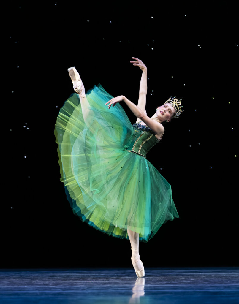 Onstage in front of a dark backdrop with stars like the night sky, Elizabeth Murphy battements her right leg forward on the effacé line while leaning back slightly, lifting her arms forward. She wears an emerald green Romantic tutu that swishes with her movement, as well as a gold crown.