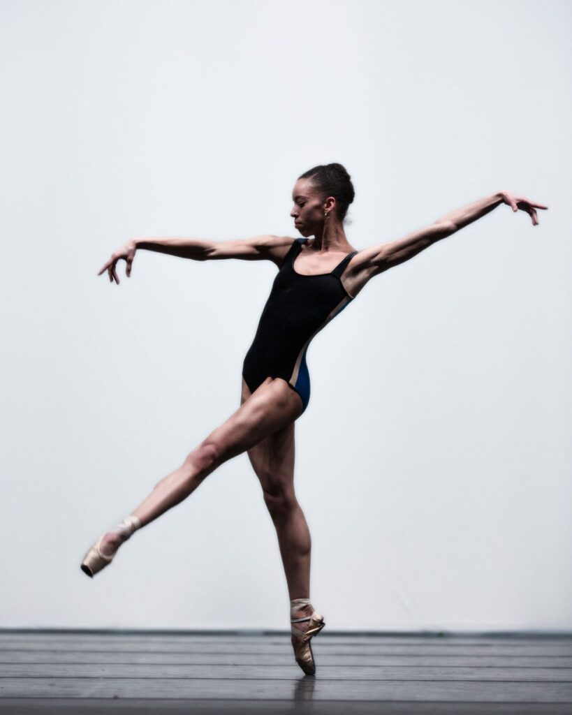 Onstage in front of a bright white backdrop, Chyrstyn Mariah Fentroy shifts her weight forward on pointe with her front leg lifted in a low croisé battement line. She extends her arms long on the diagonal to mirror her leg line, looking down slightly. She wears a black sleeveless leotard with a tan stripe down the side.