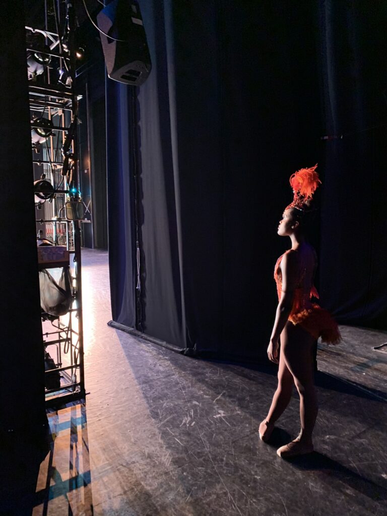 Ingrid Silva stands backstage in the wings. She wears brown tights and pointe shoes, a red costume with a short feathered tail, and red and orange feather-plumed headpiece.