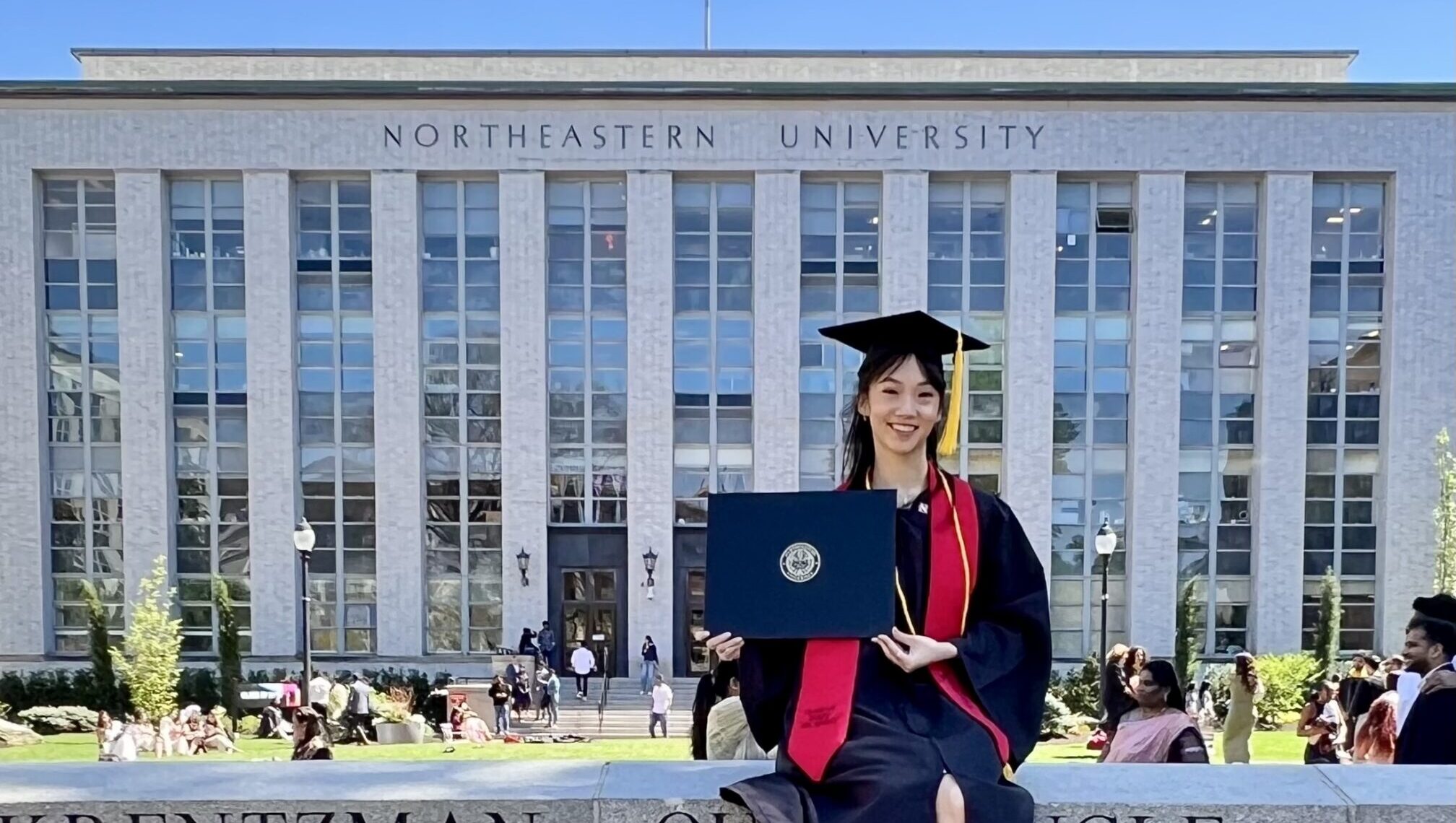 Chisako Oga, wearing a black graduation cap and gown, sits on the brick wall outside of a university building and holds her diploma, smiling for the camera.