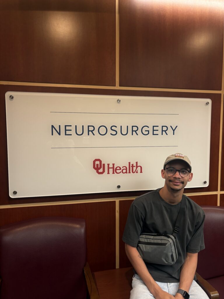 Randolph Fernandez, wearing a baseball cap, t-shirt and jeans, sits on a bench in a waiting room, front of a sign that says "Neurosurgery - OU Health."