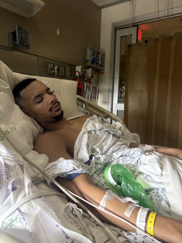 Randolph Fernandez is shown in a hospital bed with his eyes closed and various hospital bracelets and bandages taped to his right arm.