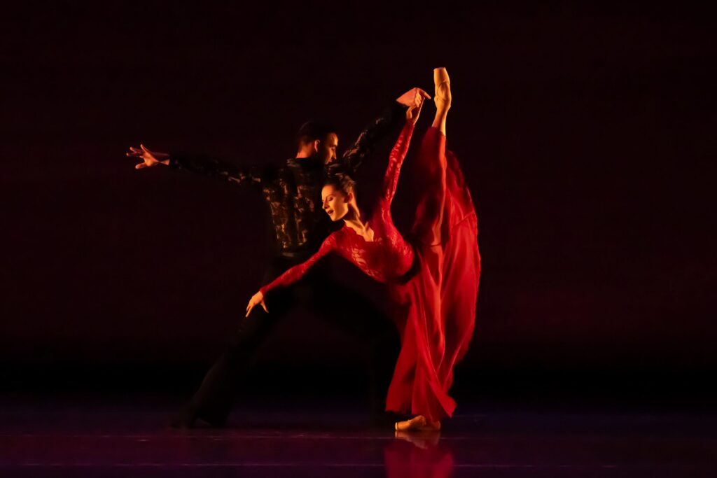 On a dark stage, Melissa Gelfin and Joshua Stayton perform a pas de deux. Gelfin, in a long red dress, penchés forward with one leg straight up in arabesque. She reaches forward with one arm and holds Stayton's hand with her other. Stayton, in a black costume with a lace back, lunges to the side facing backward as he holds her hand.
