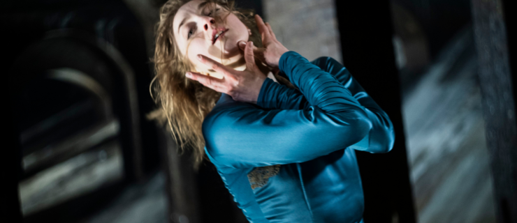 A close-up of Astrid Elbo, as Lady Macbeth, clutching her face with her arms crossed at the elbow. She throws her head back and looks at the camera with emotion, lips slightly parted. She wears a teal satin dress with long sleeves and wears her long hair down.