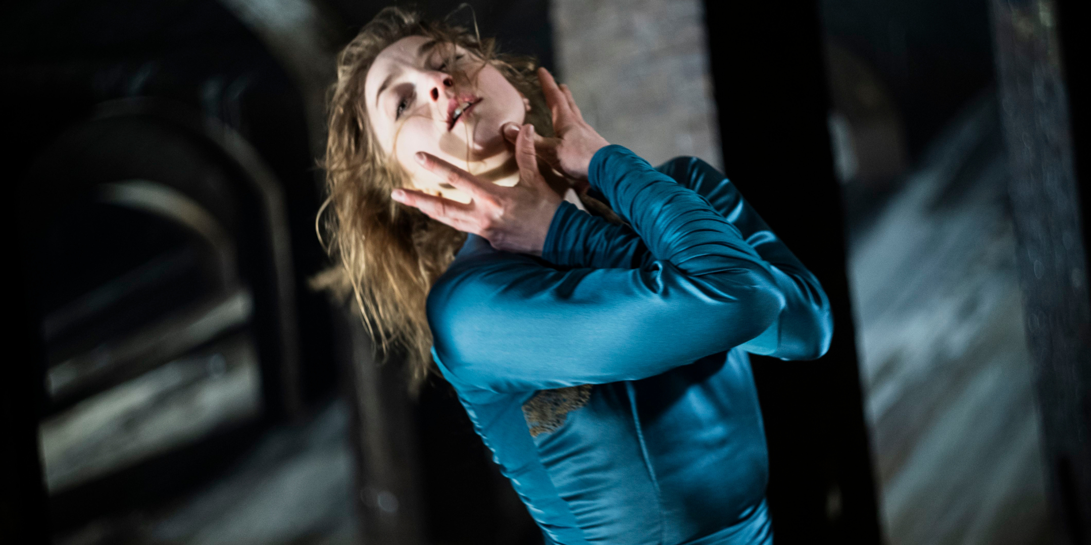 A close-up of Astrid Elbo, as Lady Macbeth, clutching her face with her arms crossed at the elbow. She throws her head back and looks at the camera with emotion, lips slightly parted. She wears a teal satin dress with long sleeves and wears her long hair down.