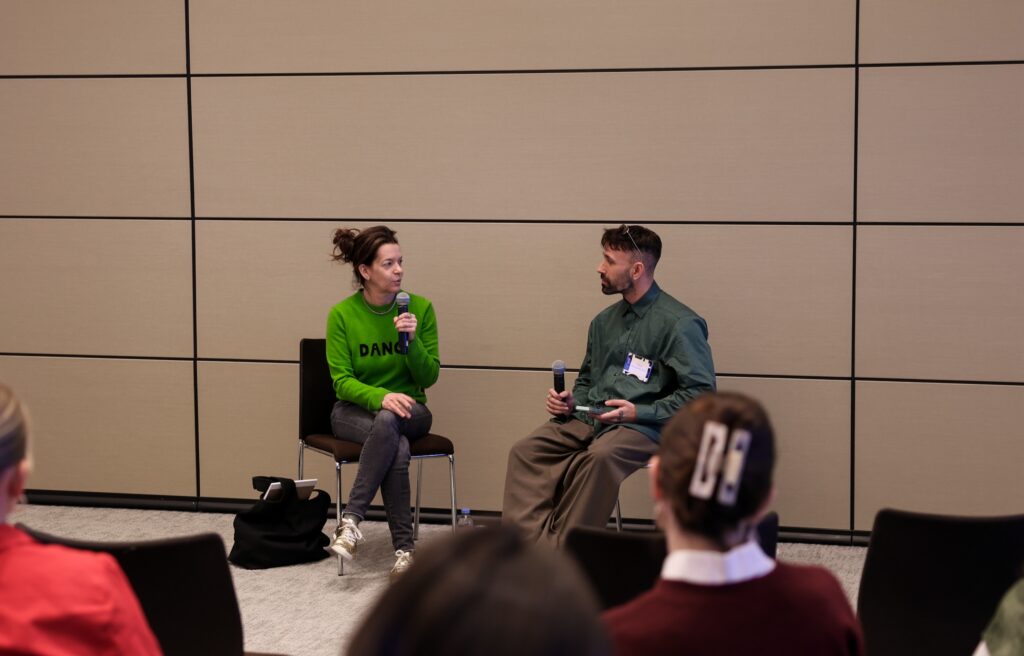 Pam Tanowitz, in a bright green sweater, and Omar Roman de Jesús, in a dark green button-down shirt, are photographed mid-conversation as they sit in chairs on a small stage.