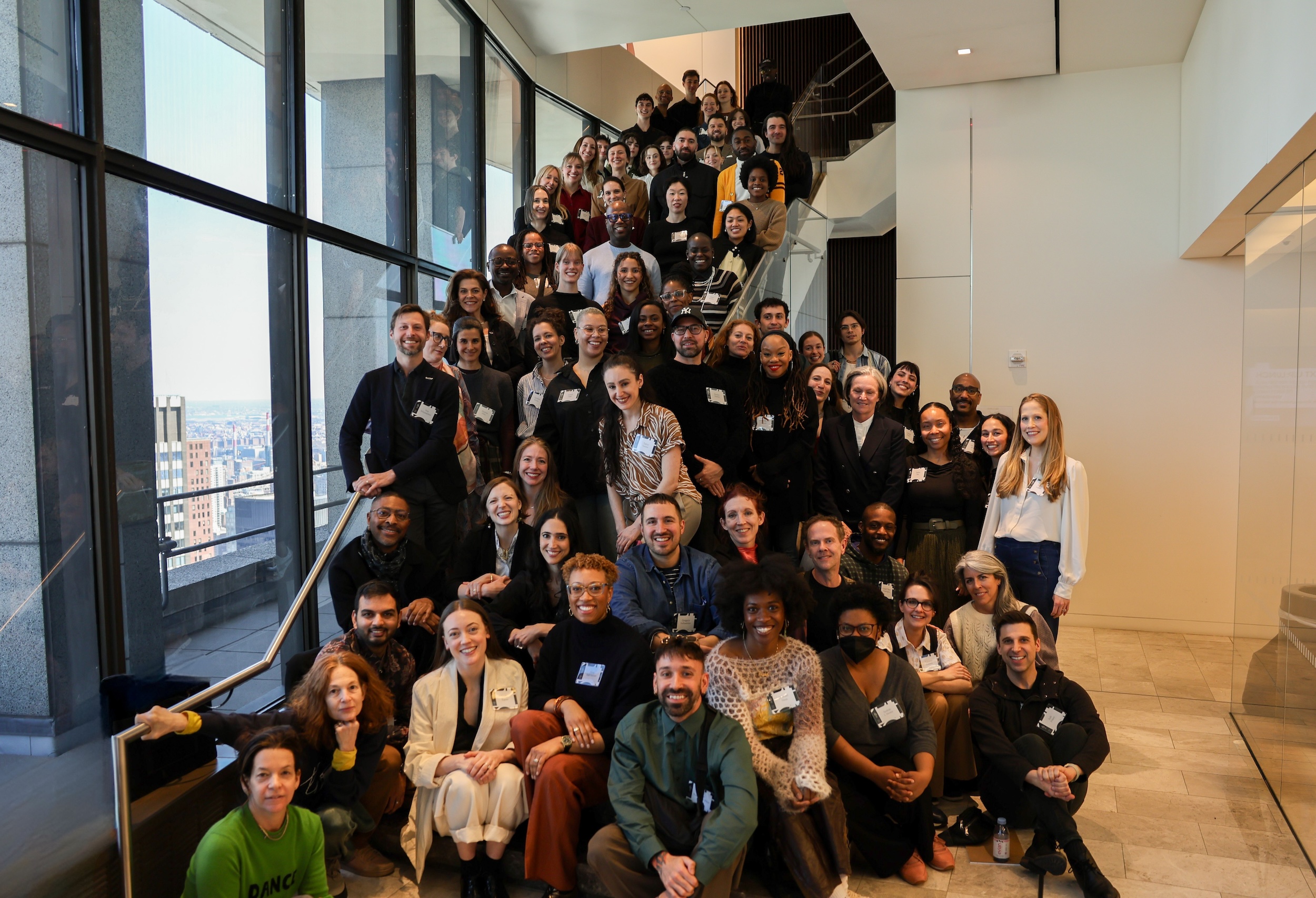 A large group of people in street clothes and nametags pose on a long curved staircase, all smiling at the camera.