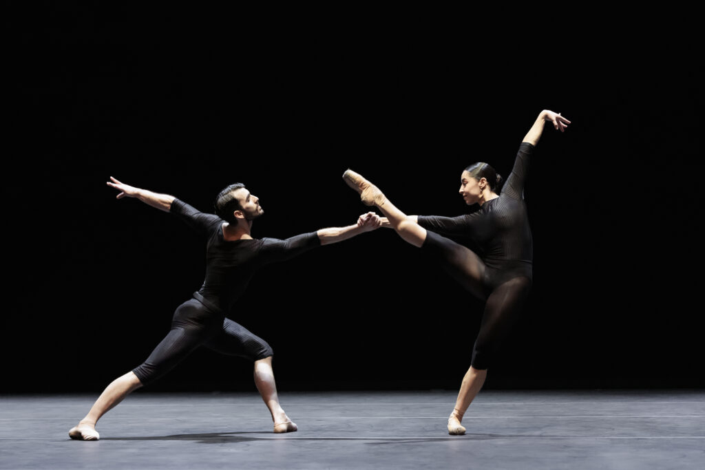 Onstage, two dancers perform a contemporary pas de deux wearing all-black. The woman, who also wears pointe shoes, plies on one leg with the other extended a la seconde. She reaches up on the high diagonal with one arm while holding her partner's hand with the other for counterbalance. He lunges forward in croisé, reaching backward with his other arm.