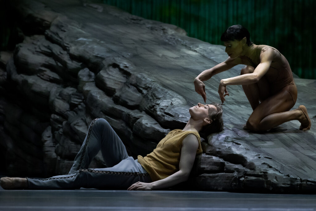 Onstage, two dancers pose on a large rock set piece. Kalle Wigle lies on his back, eyes closed, leaning back against the rock. Leroy Mokgatle crouches above him on the rock, bending forward and curving her arms with pointed fingers to gesture toward Wigle's head.