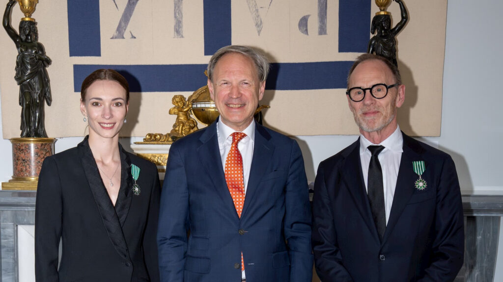 Olga Smirnova, François Alabrune, and Ted Brandsen, shown waist-up, pose side-by-side for an official photograph in front of a mantel with statues. They wear formal attire, and Smirnova and Brandsen have medals pinned to their lapels.
